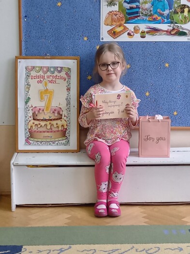 Dziewczynka siedzi na krzesełku, w rękach trzyma laurkę narysowaną przez dzieci, obok niej stoi obrazek z tortem i cyfrą 7