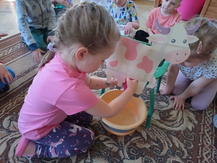Dziewczynka klęczy na dywanie, przed nią stoi krzesełko do którego przyczepiona jest papierowa ilustracja krowy, pod krowa stoi miska, dziewczynka doi krowę.