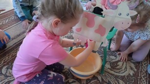 Dziewczynka klęczy na dywanie, przed nią stoi krzesełko do którego przyczepiona jest papierowa ilustracja krowy, pod krowa stoi miska, dziewczynka doi krowę.