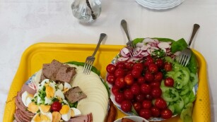 Na stole przykrytym białym obrusem stoją talerze z warzywami, wędliną, serem żółtym oraz kabanosami, na środku stoi flakon z kwiatami