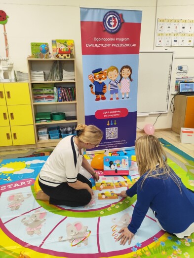 dwie kobiety siedzą na kolorowym dywanie w sali przedszkolnej, układają kolorowe obrazki, w tle banner programu Dwujęzyczny Przedszkolak