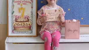 Dziewczynka siedzi na krzesełku, w rękach trzyma laurkę narysowaną przez dzieci, obok niej stoi obrazek z tortem i cyfrą 7