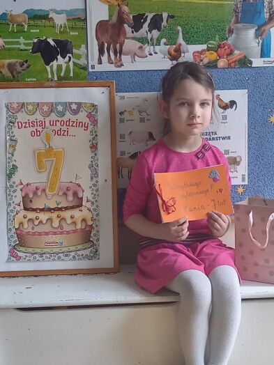 Dziewczynka siedzi na ławeczce, obok niej stoi tablica z napisem 7 urodziny, w rękach trzyma laurkę.