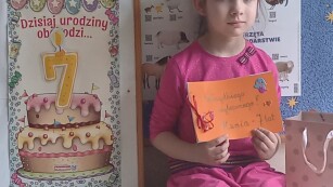 Dziewczynka siedzi na ławeczce, obok niej stoi tablica z napisem 7 urodziny, w rękach trzyma laurkę.