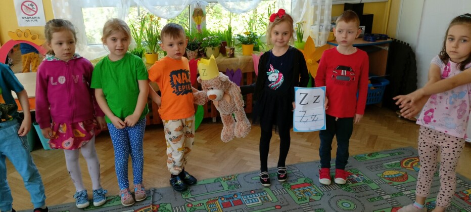 Dzieci stoją trzymając się za ręce, dzieci stojące na środku trzymają misia w koronie z literką z, oraz wydrukowaną literkę z.