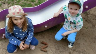 Dwoje dzieci siedzi obok zjeżdżalni na placu zabaw, obok nich leży literka z ułożona z szyszek
