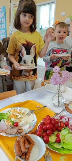 Dwoje dzieci stoi przy stole z talerzami w rękach, na stole stoi flakon z kwiatami oraz talerze z warzywami oraz wędlina i żółtym serem
