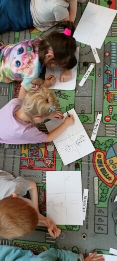 Troje dzieci leży na dywanie, przed sobą mają kartkę na której malują literkę U