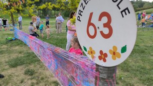 dzieci i dorośli stojący wśród zieleni, na pierwszym planie logo przedszkola 63, rozciągnięta między drzewami folia malowana farbami przez dzieci